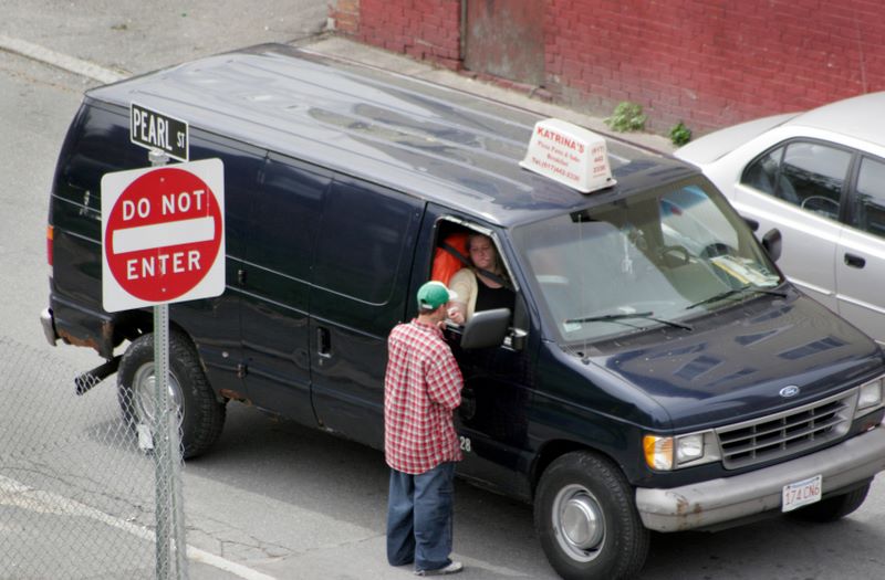 Color image of a van stopped in the road. The person in the passenger's seat leans out of the window to light the cigarette of a person standing outside of the van.