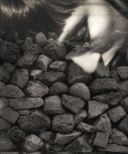 Black and white multiple exposure image of a rock wall topped with a face looking downward into the stones.