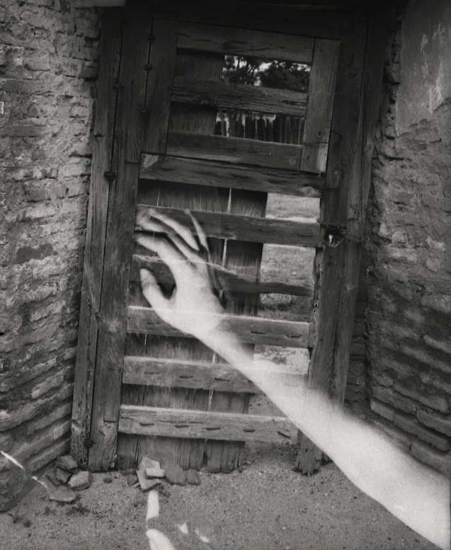 Black and white multiple exposure image of a hand reaching out to push on an old wooden door.