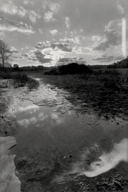 Black and white multiple exposure image of sunset over a melting lake. At the bottom of the lake in the dark water is the back side of a head, with long hair floating upward.