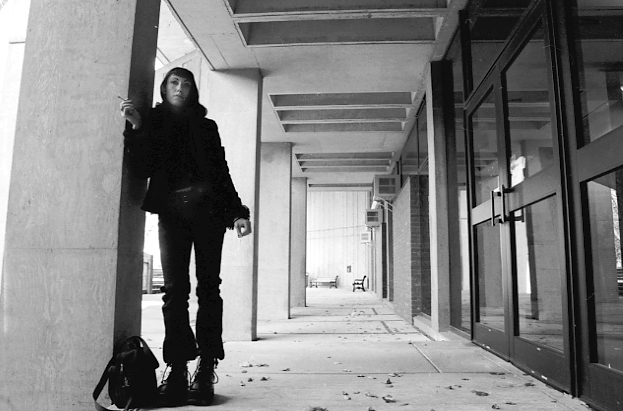 Jessica wearing all black, standing in an outdoor corridor of the UMass Lowell art department building.