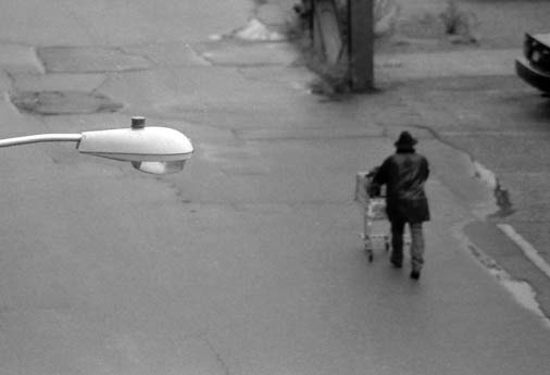 Black and white image of man pushing grocery cart down a road.