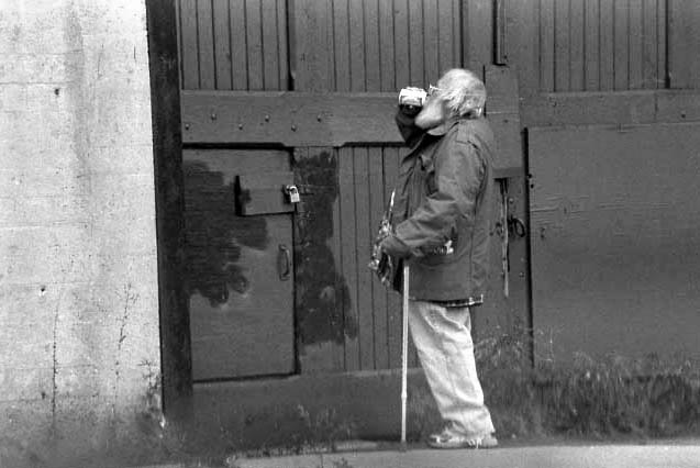 Black and white image of a man with a cane and white hair and beard, drinking from a beer can on the street.