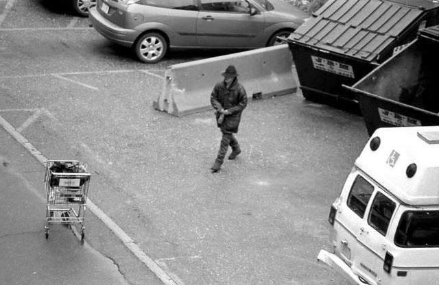 Black and white image of a man walking from a dumpster to a shopping cart, with a bottle in his hand.