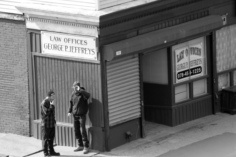 Black and white image of two men standing on a street corner smoking and talking.