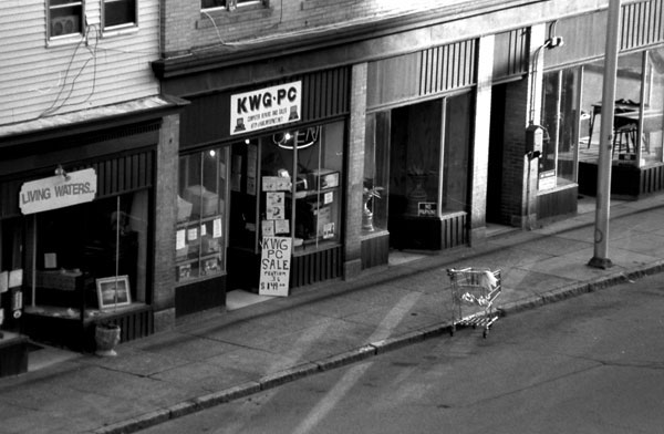 Black and white image of an empty street and storefronts, and a shopping cart in the road.