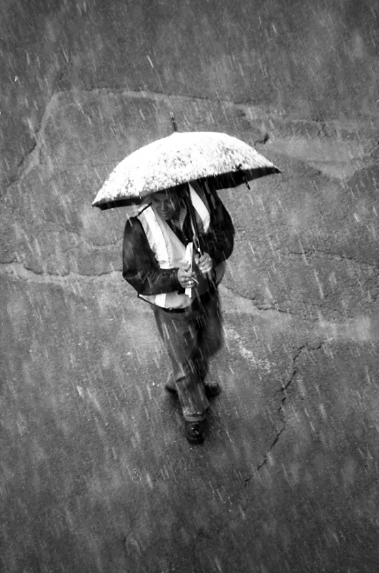 Black and white image of a crossing guard walking across the street holding an umbrella covered in snow.