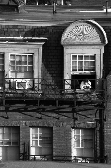 Black and white image of a person leaning out of a third story window of a residential building.