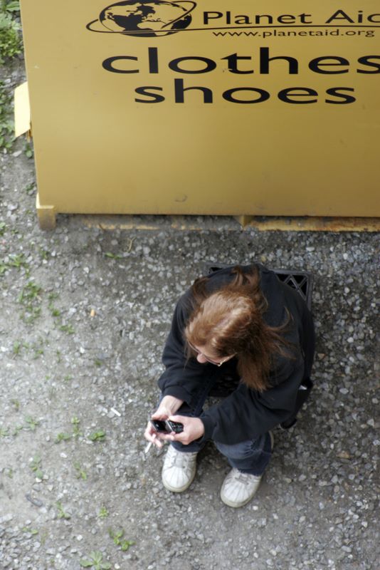 Color image of a woman sitting behind a clothing donation box, smoking and using a mobile phone.
