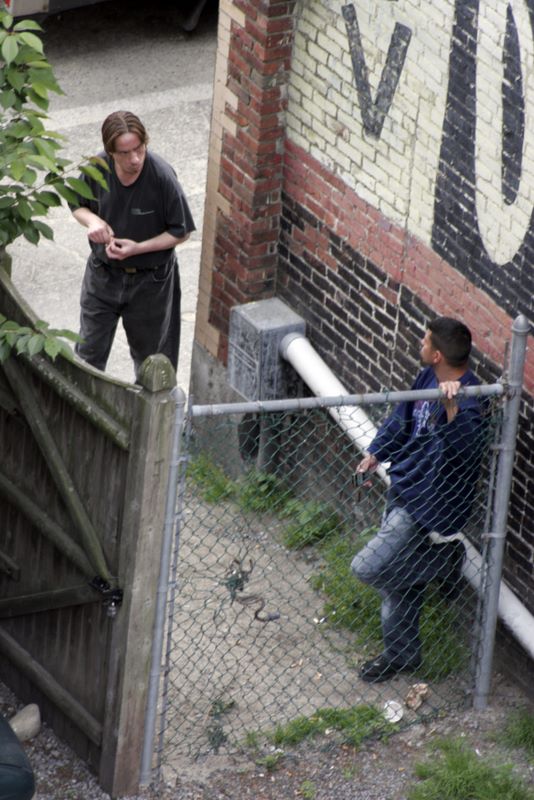 Color image of a man smoking a cigarette in a hidden alcove. Another man walking by stops and peers at him around the corner of a building.