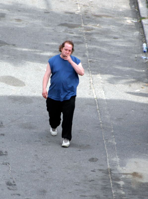 Color image of a man walking down the road smoking a cigarette.