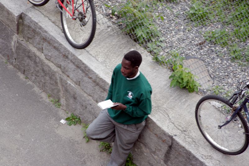 Color image of a man leaning against a low cement wall reading a book.