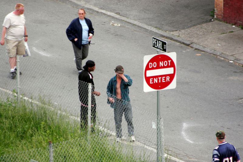 Color image of five people in the road coming from different directions. The central figure is covering his eyes with his hand, while a man next to him leans in close to say something.
